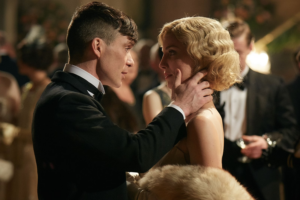 Thomas Shelby e Grace Burgess, personagens da série Peaky Blinders, estão em uma cena romântica. Thomas, vestindo um smoking preto, segura o rosto de Grace com ternura enquanto a olha nos olhos. Grace, com um vestido elegante e um cabelo loiro ondulado ao estilo dos anos 1920, retribui o olhar de forma carinhosa em um ambiente de festa, com convidados ao fundo.