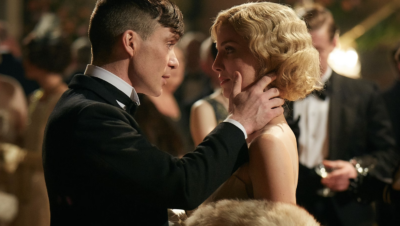 Thomas Shelby e Grace Burgess, personagens da série Peaky Blinders, estão em uma cena romântica. Thomas, vestindo um smoking preto, segura o rosto de Grace com ternura enquanto a olha nos olhos. Grace, com um vestido elegante e um cabelo loiro ondulado ao estilo dos anos 1920, retribui o olhar de forma carinhosa em um ambiente de festa, com convidados ao fundo.